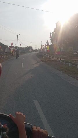 This video shows a point-of-view shot from a moving vehicle, likely a motorcycle, traveling along a paved road. The road is lined with a variety of flags, suggesting a festive or celebratory event. These flags are colorful, featuring red, white, yellow, and green, and are attached to poles along the side of the road. Some buildings are visible in the background, and the overall scene suggests a rural or semi-rural setting in Dente Makmur, Indonesia. The sun is low in the sky, indicating either early morning or late afternoon, and the light is bright and somewhat hazy, suggesting a warm, possibly humid, day. The activity taking place is travel along the road, with other vehicles, including at least one motorcycle, visible ahead. The focus is on the journey and the decorated roadside. The hands of the person holding the camera are visible in the foreground, gripping a handle or steering mechanism. There is no visible text in the video.