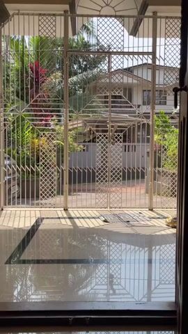 The video opens with a view through an open doorway to a gated entryway. The gate is made of white metal bars and features a decorative diamond pattern. Sunlight casts shadows on the tiled floor of the entryway. Beyond the gate, a street is visible with houses and lush green foliage. Cars are parked in front of one of the houses. The video then zooms in slightly on the gate, showing a lock mechanism. The view pulls back to reveal the doorway more fully, with a wooden door on the right side of the frame. The room beyond the doorway appears to be an interior space with a ceiling fan visible. The video continues to show the exterior view through the open doorway and the gate. The overall impression is of a residential property with an emphasis on security and a tropical outdoor environment.