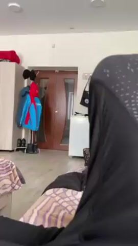 A person in a black hijab sits on a bed in a room. The bed has a patterned bedspread. Behind the person are two beds, a wardrobe, a coat rack with coats, shoes, and a red hat, a dark wood door, and a small white refrigerator. The room is well-lit.