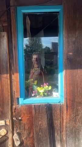 This media file shows a reflection in a wooden window pane. The window frame is painted a bright blue. A young woman is visible in the reflection, holding a smartphone and taking a selfie. She is wearing a colorful, patterned dress. In the foreground of the reflection, on the windowsill, is a small bouquet of flowers, primarily yellow and pink.

Behind the woman, the reflection shows a rural scene. There are trees, buildings that appear to be sheds or small houses, and a cloudy sky. The overall impression is one of a sunny day, likely in the afternoon, given the lighting. The wooden exterior of the building suggests a rustic or older structure. The setting appears to be outdoors, possibly a yard or garden area. There is no visible text in the media file. The woman's expression is neutral, focused on taking the photograph.