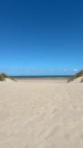 The video shows a view of a sandy beach with the ocean and sky in the background. The camera is positioned at eye level, looking out towards the water. In the foreground, there is a expanse of light-colored sand with some ripples and footprints. To the left and right, dunes covered in dry grass are visible. In the middle ground, a stretch of wet sand or a tidal flat is apparent, with some small figures of people walking and yellow buoys scattered across it. Beyond the beach, the sea is a dark green color, meeting a clear blue sky dotted with a few white clouds. Sailboats are visible on the horizon. The overall impression is a sunny day at the beach.