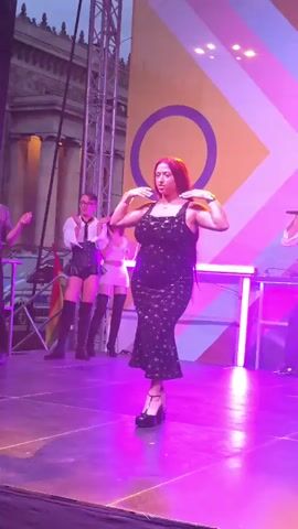 A woman with long red hair in a black floral dress and platform heels dances on a stage. She is holding a microphone and appears to be singing. Behind her, a man in a white shirt and suspenders also holds a microphone and sings. To the left of the woman, two individuals in white shirts and skirts with thigh-high boots stand and clap. In the background, a large banner displays a geometric pattern with the colors yellow, pink, and light blue, and a large purple circle. The stage is lit with purple and green lights.