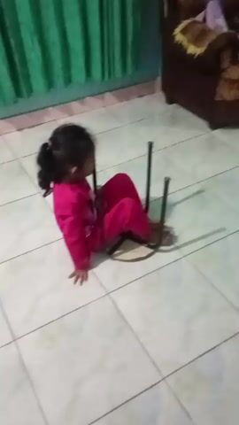 A young child wearing a pink outfit sits on a tiled floor. Another child, wearing a blue shirt and tan pants, kneels on the floor while holding onto a metal structure that appears to be part of a chair. The second child pushes the metal structure, moving it forward. The first child reaches out a hand toward the second child. A brown couch is visible in the background.