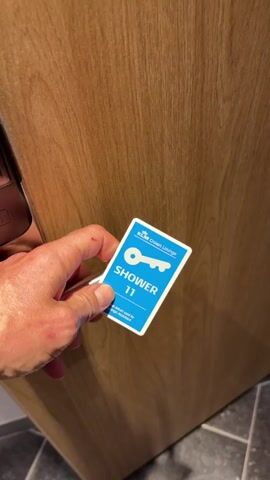 A hand holds a blue key card with a picture of a key and the word "SHOWER 11" on it. The card is in front of a wooden door with a metal handle. The scene then shifts to a bathroom with a sink, a mirror, and a stool. The person taking the video is reflected in the mirror, wearing a t-shirt, shorts, a cap, and headphones. The video then shows the interior of a shower stall with white walls.