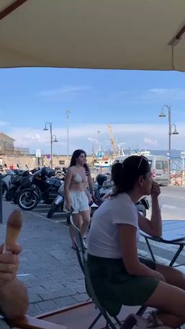 The video depicts a scene in Porto Santo Stefano, Italy, on a sunny day. The camera is positioned under a large umbrella, looking out towards a harbor. In the foreground, two women are seated at a small outdoor table, seemingly at a cafe. The woman on the left is wearing a white t-shirt and a green skirt, while the woman on the right is partially visible, wearing a white top and a dark skirt. 

In the middle ground, a young woman in a light-colored bikini and white shorts is standing near a parked scooter, looking towards her phone. Another young woman, also in a bikini and shorts, is walking past her, holding an ice cream cone. Behind them, a row of parked scooters and motorcycles are visible along a paved walkway. 

The background reveals the picturesque harbor of Porto Santo Stefano, with numerous boats and yachts docked. A crane is visible in the distance, suggesting ongoing port activity. The shoreline is lined with buildings, and a lush, green hillside covered in houses rises behind the town. The sky is a clear blue with a few scattered clouds, indicating fair weather. The overall atmosphere appears relaxed and summery, typical of a coastal Italian town.

The only visible text is on a white bag attached to the back of a scooter, which reads "K-UV". The time of day appears to be late morning or early afternoon, given the bright sunlight and shadows. The emotions conveyed are casual and leisurely, with people enjoying the seaside atmosphere.