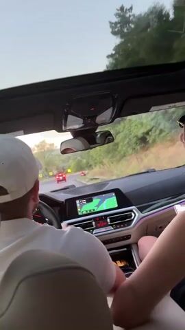 The video shows the interior of a car from the perspective of the back seat, looking forward. Two people are visible inside the car. The driver, a man wearing a white baseball cap and white shirt, is steering the vehicle. The front passenger's shoulder and hair are visible. The car has a large infotainment screen displaying a navigation map. The car is driving on a paved road that curves through a wooded, hilly area. The sun appears to be setting, casting a warm light. In front of the car, two other vehicles, one red and one dark-colored, are visible on the road ahead. The sunroof of the car is open, allowing a view of the sky and trees.
