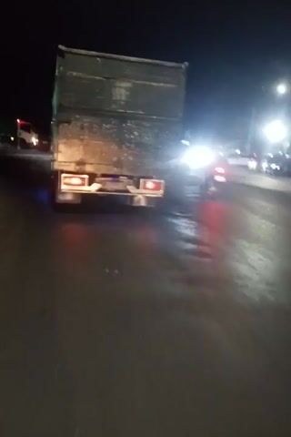 The video shows a truck with a green cargo container being driven at night on a wet road. The truck has its brake lights and hazard lights illuminated, and there are also blue and red lights on the back. The road surface appears to be asphalt and is reflecting the lights from the truck and nearby streetlights. There are other vehicles and buildings visible in the background. The video appears to be filmed from another vehicle traveling behind the truck.