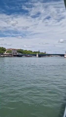 The video presents a serene daytime view of a cityscape along a wide river, identified as Lyon, France. The camera slowly pans from left to right, offering a panoramic glimpse of the urban environment from what appears to be a moving boat on the river.

The sky overhead is a vibrant blue, adorned with numerous fluffy white clouds that suggest pleasant weather conditions. In the foreground, the river's surface is visible, showing gentle ripples that indicate movement, likely from the vessel filming the scene.

Across the river, the city's architecture is a prominent feature. Classic European-style buildings, multi-storied with light-colored facades, line the riverbank. Many of these structures exhibit typical architectural details, including visible chimneys. A continuous stretch of lush green trees forms a natural border between the water and the buildings, adding to the picturesque quality of the scene.

Initially, a long, multi-arched bridge spans the river in the middle ground, connecting the city's banks. Several boats, possibly barges or smaller river cruise vessels, are visible near the far bank, either moored or navigating the waters slowly. As the camera continues its smooth pan to the right, the bridge gradually moves out of frame, revealing more of the charming urban landscape and the continuous line of historical buildings along the waterfront.

The video captures a peaceful moment, highlighting the architectural beauty of Lyon against a backdrop of clear skies and calm waters. There are no discernible people or visible text within the frame.