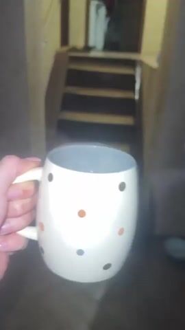 A hand holds a steaming mug with a light-colored body decorated with multi-colored dots. The mug is filled with a dark liquid, likely coffee or tea. The background shifts from wooden stairs to a room with tiled flooring. The steam from the beverage is visible. No faces are visible.