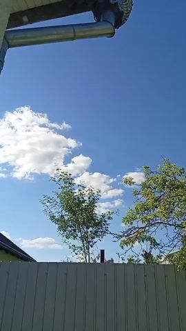 The video captures a clear blue sky with scattered white clouds. In the foreground, a grey metal fence runs horizontally across the lower part of the frame. Above the fence, green trees with dense foliage are visible. A metal downspout extends from the upper left corner of the frame. There are no people or animals visible in the video. The scene appears to be an outdoor setting on a sunny day.