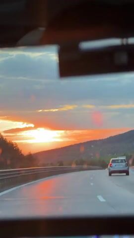 The view from inside a vehicle moving along a highway shows a vibrant sunset. The sun is partially obscured by clouds and a distant treeline. A metal guardrail runs along the side of the road. The interior of the vehicle is partially visible in the foreground, providing context for the viewer's perspective.