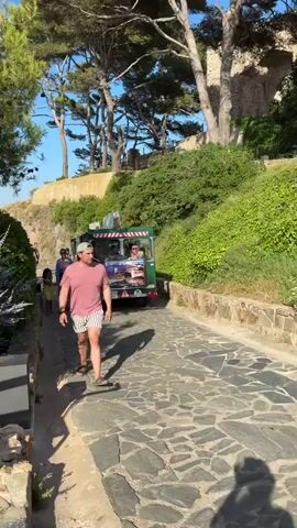The video depicts a sunny day in a coastal town, likely Tossa de Mar, Spain, as suggested by the scenery. A man in a pink t-shirt and striped shorts walks down a cobbled path, his shadow stretching before him. He wears a light-colored baseball cap and sandals. Behind him, a small green tour bus, with people visible inside and on the roof, follows the path. Two women are walking ahead of the man, towards the bus.

The path is bordered by lush green vegetation and stone walls. Tall trees with pine-like needles provide shade. In the background, remnants of stone structures are visible on a hillside, suggesting historical significance or ruins. The sky is a clear blue, indicating pleasant weather.

Further along the path, a metal trash can and an informational sign are visible. The sign features text and images, though they are too small to be legible in detail. The overall atmosphere is relaxed and leisurely, consistent with a tourist location on a warm day. The presence of the tour bus and people exploring the area suggests a guided tour or a visit to a scenic viewpoint.