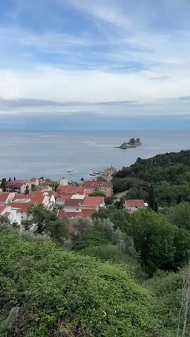 The video shows a panoramic view of a coastal town with red-tiled roofs nestled between green hills and the sea. The sea is calm and blue, with a small island visible in the distance on the right and larger landmasses forming the horizon on both sides. The foreground is dominated by dense green vegetation. The sky is blue with scattered clouds. The camera pans slowly to the left, revealing more of the coastline.