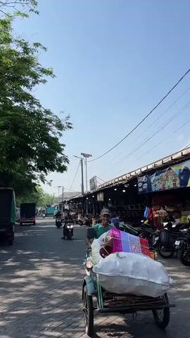 A motorcycle rider, an elderly man wearing a green t-shirt and a cap, is in the foreground, smoking a cigarette and looking towards the camera. He is on a motorcycle laden with goods, including a pink basket. In the background, a street scene unfolds with other motorcycles, a truck, and a row of market stalls under a clear blue sky. The market stalls appear to be selling various items, some of which are visible as colorful fabrics and other merchandise. Trees line the left side of the street, providing shade. The overall atmosphere is that of a bustling outdoor market on a sunny day.