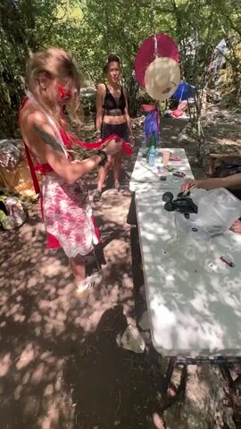 The video shows a gathering of people in a wooded area. In the foreground, a person with blonde hair wearing a red top and a white and pink skirt is holding a red ribbon. To their left, a person with dark hair and a black top and shorts stands looking towards the camera. To the right of them, a white folding table is set up with various objects on it, including bottles, containers, and what appears to be a small remote-controlled car. Further back, a person with blonde hair and headphones is sitting at the table. The environment is a forest with trees, sunlight filtering through the leaves, and the ground covered in dirt and leaves. There are also some camping-like structures visible in the background.