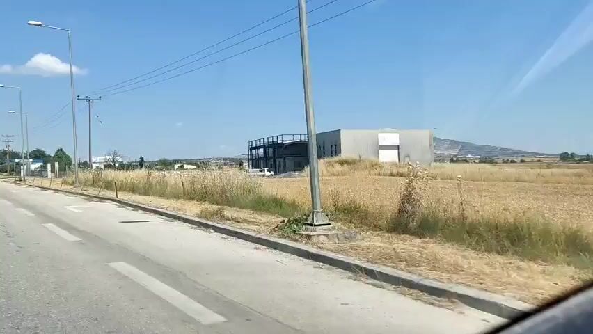 A dashcam view from a moving vehicle shows a rural landscape on a sunny day. A two-lane road with a painted white arrow on the asphalt occupies the left side of the frame. Utility poles with power lines run alongside the road. A metal fence separates the road from a dry, grassy field. In the distance, a gray building with a white loading dock is visible, next to a darker building with an exposed metal frame that appears to be under construction. Beyond the buildings, more fields and distant hills under a clear blue sky with some scattered clouds can be seen. The vehicle is moving, and the view is shifting to the right.