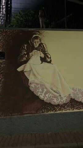 The video shows a mural on a wall. The mural depicts, from left to right, an elderly woman sewing a piece of cloth, a man leaning over a basket, and two more men standing. The mural is painted in a sepia tone, and the wall is beige. The video pans from left to right, showing the entire mural. The background includes a black fence, palm trees, and a dark sky. The video is shot at night.
