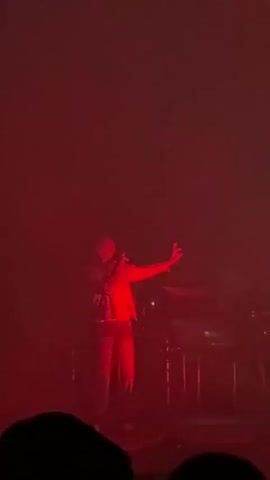 The video shows a dimly lit concert or performance space illuminated by vibrant red lights. In the center of the frame, a person, likely a performer, is visible from the back, wearing a light-colored jacket and dark pants. They are standing and gesturing with their right arm extended and pointing forward. Behind the performer, other figures are partially obscured by shadow and the red lighting, possibly other performers or band members, and some musical equipment such as keyboards or consoles. The foreground shows the silhouetted heads and shoulders of audience members, indicating a live event. Smoke or haze is present in the air, contributing to the atmospheric lighting. The overall impression is of an energetic and immersive performance.