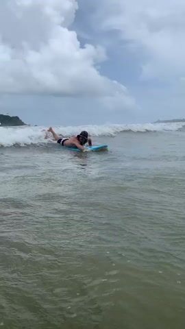 A man is shown surfing in the ocean. He is on a blue surfboard, paddling with his hands. The water is choppy with white waves. In the background, there is a shoreline with trees and a cloudy sky.