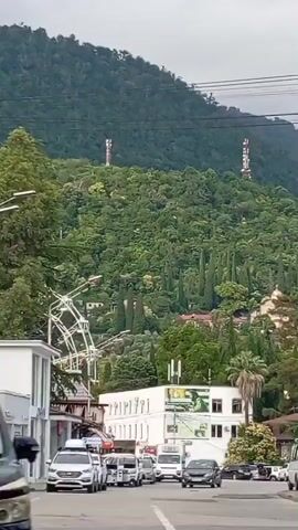 The video pans across a street scene with a lush green hillside in the background. Several cars are visible on the road, driving in the same direction. On the hillside, there are buildings and tall trees, including cypress trees. Two communication towers with red and white banding are also visible on the upper part of the hillside. In the foreground, a large tree with light-colored blossoms is prominent. To the right of the tree, an unusual building with a distinctive, curved roof covered in colorful mosaic tiles is partially visible. The sky is overcast.