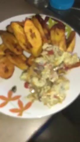 A plate of food is shown on a kitchen counter. The plate is white with a floral design around the edge. On the plate are fried plantains and scrambled eggs. The plantains are golden brown and cut into diagonal slices. The scrambled eggs contain red and white pieces. In the background, there is a clear container and a blue container. The video is initially out of focus but becomes more focused on the plate of food.