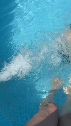 The video shows a person's legs and feet submerged in a bright blue swimming pool, with water splashing around them. In the background, another person wearing a black swimsuit and cap is swimming, creating a trail of bubbles and ripples. The camera appears to be positioned close to the water's surface, looking down at the scene.