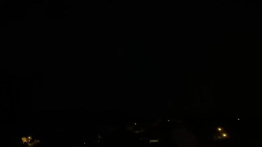 The video is a nighttime shot of a dark sky over a dimly lit landscape. There are scattered lights that appear to be from streetlights or houses, suggesting a rural or suburban area. The sky is predominantly black, but there are brief, faint flashes of light, possibly indicating distant lightning or some atmospheric phenomenon. No people or discernible objects are visible in the foreground or midground, with the focus being on the vast, dark expanse above and the faint terrestrial lights below. The video's duration is very short, and the lighting conditions are low, making it difficult to discern fine details.