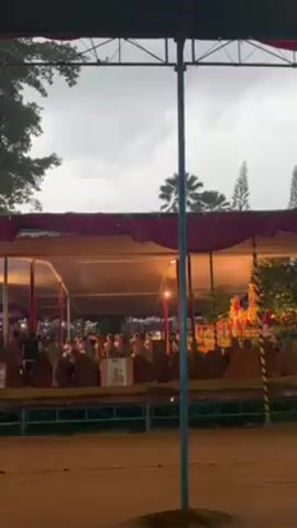Okay, here's a detailed description of the video:

The video captures an outdoor performance taking place under a large, partially covered stage or tent. A group of people, who appear to be monks based on their robes, are seated in rows, facing the stage. On the stage, a group of performers, possibly dancers or actors, are visible. They are standing in front of the seated monks.

The setting seems to be outdoors, indicated by the presence of trees in the background, including palm trees. The sky is overcast and gray, with a slight drizzle or rain falling, which can be seen in the foreground of the shot. A tall pole stands prominently in the foreground, partially obscuring the view.

The stage area is illuminated with artificial light, creating a brighter area under the canopy, contrasting with the darker outdoor environment. People standing in the audience can be seen beyond the line of monks.

The overall atmosphere suggests a cultural event or performance, possibly of religious or ceremonial significance.