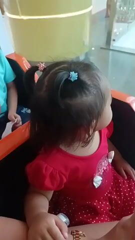The video shows a group of children in what appears to be a carousel or amusement ride. In the foreground, a young girl wearing a red dress with white polka dots is facing away from the camera. To her left, a boy in a light blue t-shirt with a monster graphic is looking to the side. Further back, a girl with pigtails is leaning back and smiling at the boy. The background shows a brightly lit indoor environment, possibly a mall, with decorative elements and other people in the distance. The ride itself is a dark-colored vehicle with orange trim.