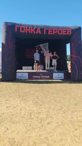 Four young people stand on a podium under a large inflatable archway that reads "GONKA GEROEV" in Russian. The archway is black with red contour lines and has a red banner inside that says "SOVET POPOSTOV". The podium has three tiers, with the middle tier labeled "2" and the top tier occupied by two people. The person on the left of the middle tier wears a blue t-shirt and shorts and is holding a flag. The person in the center of the top tier wears a pink t-shirt and light-colored shorts and is waving. Another person in a black t-shirt and shorts stands to the left of the person in the blue t-shirt. A fourth person in a black t-shirt and shorts stands to the right of the person in the pink t-shirt. The podium is decorated with logos, including "GAZPROM BANK" and "GONKA GEROEV". The ground in front of the podium is covered in sand and grass, and the sky is clear and blue.
