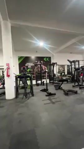 The video shows a person using a leg extension machine at a gym. The camera is positioned above the person's legs, showing their white sneakers, bare thighs, and the machine's black padded bar. The gym is brightly lit with gray floors and various weight machines in the background, most in shades of black and green. A motivational poster is visible on the back wall. The person is performing leg extensions.