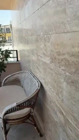 The video shows an outdoor patio. The patio has a settee and a small round table. A large potted plant sits between the settee and table. The patio has beige tile flooring. The patio wall is made of beige stone. The patio has a black metal railing that runs around the perimeter. In the background, buildings and blue skies are visible.
