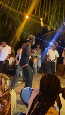A video captures an outdoor nighttime gathering of approximately a dozen people, illuminated by festive blue and yellow lights strung beneath palm fronds.  Several individuals are standing and observing a male dancer in a black tank top and pants, who performs energetic movements on a sandy surface.  Attendees, dressed in casual attire including white shirts and khaki trousers, are seen holding phones, presumably recording the performance.  A few seated spectators are visible in the foreground, their backs to the camera.  A blue plastic chair is positioned near the dancer. The overall setting suggests a celebratory beach party.