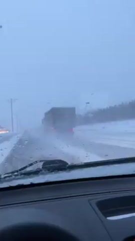 The video shows a view from inside a car driving on a snowy highway. A large dump truck is visible ahead in the same lane, kicking up snow and water. The windshield wipers are active, clearing the snow and water from the windshield. Trees line the side of the road, also covered in snow. Other vehicles are visible in the distance. The lighting is flat due to the overcast snowy conditions.