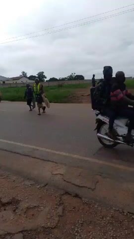 A group of people are walking on a paved road under an overcast sky. They are moving from left to right across the frame. A vehicle is visible on the left side of the road. There is a grassy embankment on the right side of the road with a few people standing or walking on it. Buildings and structures are visible in the background. The road surface appears to be damaged in places.