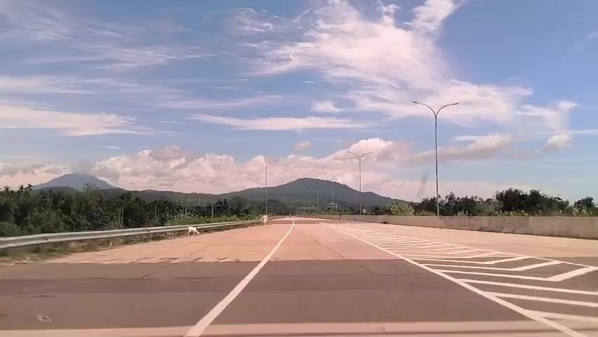 The video shows a forward-facing view from a vehicle driving on a four-lane road. The sky is blue with scattered clouds. Mountains are visible in the distance.  Streetlights line the road. A white dog is briefly visible on the left side of the road near the shoulder.