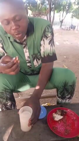A man is sitting on a bench at a wooden table outside under trees. He is wearing a matching green outfit patterned with black palm trees and white trim. He is holding a small piece of food in his left hand, and he is drinking from a white plastic cup with his right hand. On the table are a red plate with food scraps, and a small blue plate. In the background is a dirt ground with other picnic tables under the trees. There is a woman in pink walking in the background. The sun is shining brightly.