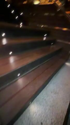 The video shows the exterior of a restaurant named CABANA RESTAURANT & COCKTAIL BAR at night. The entrance features a wide, dimly lit staircase made of dark wood with small lights embedded in the risers. A metal railing lines the right side of the stairs. The restaurant itself is brightly illuminated with warm, orange lights. The building has a modern design with angular canopies and textured walls. Plants are visible in planters near the entrance. The overall ambiance suggests a lively and upscale dining or bar establishment.