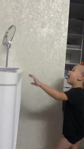 This media file captures a playful interaction between two young children in what appears to be an indoor setting, likely a home in Нижнеяркеево, Russia. The primary subjects are two boys, both wearing black t-shirts and dark shorts. The first boy, positioned more centrally and in focus for part of the clip, is actively engaged in reaching towards a small, round mirror on a stand. He exhibits clear joy and excitement, with his mouth open in a laugh or shout.

The second boy, partially visible and slightly out of focus, is interacting with the first boy, his hands on his back, suggesting a game of tag or a playful chase. The setting includes a light-colored, textured wall and a white cabinet or dresser. In the background, a tall shelving unit is visible, containing various items, and a doorway or hallway leading to a darker area. The flooring is a light wood laminate.

The overall activity is one of energetic play and sibling interaction, characterized by laughter and movement. The lighting suggests it is daytime, as there is no indication of artificial lighting being the primary source, and the overall ambiance is bright and airy. The emotions conveyed are overwhelmingly positive, centered around fun and shared amusement. There is no visible text in the media file.