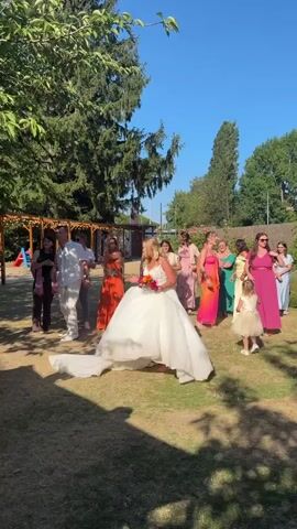 The video shows a wedding reception outdoors on a sunny day. A bride in a large white wedding dress with a bouquet of colorful flowers is seated on a lawn. She is surrounded by a group of women, most of whom are dressed in colorful attire. A man in a light-colored suit walks away from the bride, towards a group of people who appear to be guests. The guests are clapping and cheering for the couple. The environment is a grassy area with trees and a wooden pergola structure in the background. The overall atmosphere appears to be festive and celebratory.