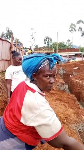 The video shows a woman looking at the camera while standing on the edge of a large trench dug into reddish-brown soil. The trench is filled with water. In the background, other individuals are working in the trench, passing buckets and containers between them. One person wears a red hard hat and a yellow safety vest, while others wear hats and yellow shirts. The overall environment appears to be a construction site or area where excavation is taking place.