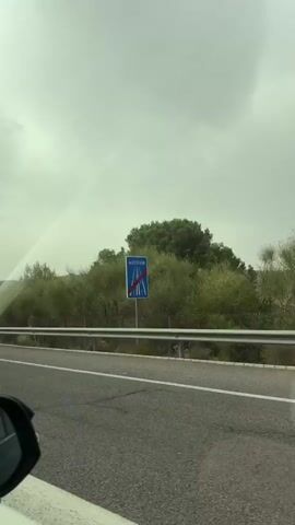 The video shows a view from a car driving on a road. The road has white lane markings. A metal guardrail runs alongside the road. Beyond the guardrail, there is vegetation, including bushes and trees. In the background, there are hills. The sky is overcast, suggesting it is daytime and potentially cloudy or hazy weather. There are road signs visible, including a sign with a "9" on it, a sign with black and white chevron arrows, and a yield sign with a red triangle. A green and white striped traffic bollard is also visible. The car appears to be in a rural or semi-rural setting, likely in Lorca, Spain, based on the provided location context. The mood is neutral, as it is a straightforward recording of a drive. There is no explicit indication of emotion.