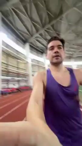 A man wearing a purple tank top and black shorts runs on a red indoor track with white lines marking the lanes. He is holding the camera in front of him at arm's length, capturing a selfie perspective as he runs. The background shows a green turf field next to the track, with netting separating the two. The building has a high, light-colored ceiling supported by visible beams.