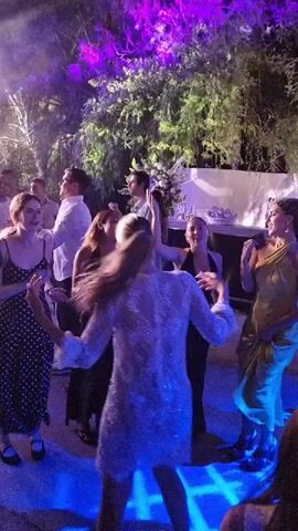 A group of people are dancing outdoors at night. Purple and white lights illuminate the scene, casting beams through trees and creating an atmospheric party environment. Several women in dresses are visible, some with their arms raised and moving to the music. Men in white shirts are also dancing among them. A table with bottles and glasses is visible in the background, suggesting a celebration. The overall mood appears festive and lively.