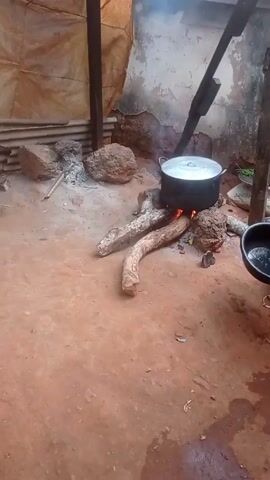 The scene appears to be an outdoor cooking area, possibly in a rural setting. A large metal pot sits atop a wood fire, supported by rocks and thick branches. Smoke rises from the pot. To the left, a green bucket sits on the reddish-brown ground, alongside a bottle of what appears to be cooking oil, a small bag, and a water bottle. Rocks and corrugated metal sheets are visible in the background, near a makeshift shelter created with wooden beams and a tarp-like material.  The overall environment looks simple and functional.