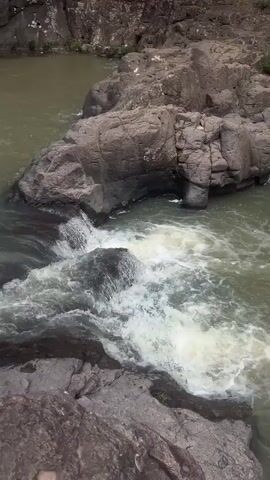 The video shows a waterfall cascading down a rocky cliff face into a body of water. The water is murky green, and the rocks are dark and weathered. Lush green vegetation grows on the cliffs above the waterfall. The video pans up the waterfall, showing its different tiers and the surrounding landscape. In the foreground, a person's legs are visible, submerged in the water. The sky above is a mix of blue and white clouds. The sound of rushing water is present throughout the video.