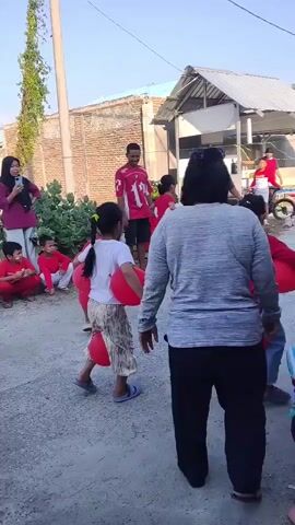 The video shows an outdoor event, possibly a children's party or competition, taking place on a gravel path between buildings. Several children, mostly wearing red shirts and pants, are participating in a game involving balloons. One child, in a white shirt and patterned pants, is crouched with two red balloons wedged between their legs. Other children are also seen with balloons between their legs. Adults are present, some observing and others appearing to be supervising the activities. One man in a red shirt with the number 12 on the back is standing and holding a red bag. Another man in a dark shirt and hat is also standing and observing. The environment appears to be a residential area with simple buildings made of brick and corrugated metal. The lighting suggests it is daytime, with a bright sky. The overall atmosphere seems to be one of casual fun and community gathering.