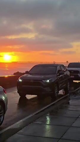 A drone's eye view shows a sunrise over a body of water, with a pier visible in the distance. Cars are parked along a wet, paved area next to the water. The sky is filled with clouds reflecting the orange and yellow hues of the rising sun. Waves are crashing against a barrier along the edge of the water, and spray is visible in the air. The scene is illuminated by the soft light of dawn.