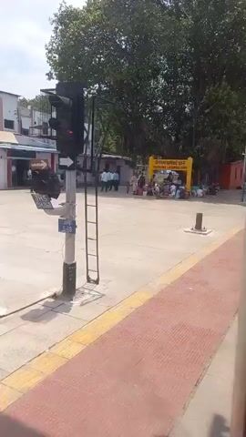 The video shows the railway station named "Virangana Lakshmibai Jhansi". A group of people are sitting on the platform near the station sign. Some people are walking around, and others are sitting on blankets. There are bags and luggage scattered around the people on the ground. The day is bright and sunny, with a clear sky visible in the background. A ladder is visible. A tree is in the background.