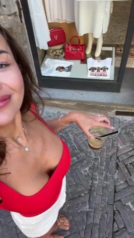 This video captures a woman in a red top and white skirt holding a coffee cup, walking through a European-style town square. The woman is smiling at the camera, suggesting a positive and leisurely mood. She is wearing a silver necklace with a square pendant and a bracelet. In the background, a storefront window displays handbags and clothing, hinting at a shopping district.

The scene transitions to a street with cobblestone pavement and old buildings, one of which has a sign reading "TRATTORIA." A man is seen taking a photo with his phone, and further down the street, a musician plays a violin. People are walking, sitting at outdoor cafes, and enjoying the day. The architecture and the presence of a trattoria suggest a location in Italy, confirmed by the prompt as Mozzo, Italy.

The video culminates in a wider view of the square, featuring a stone fountain surrounded by people sitting at tables under umbrellas. The sunlight indicates it is daytime, likely late morning or afternoon. The overall atmosphere is one of a pleasant day in a charming Italian town, with people engaged in various activities like shopping, dining, and enjoying the street life. There are no other distinct text elements visible beyond the "TRATTORIA" sign.